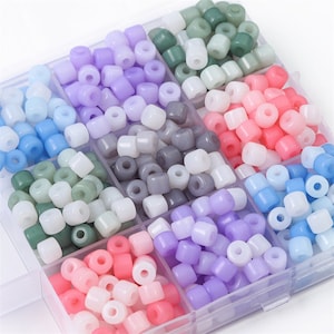 Könnte beinhalten: Eine transparente Plastik-Aufbewahrungsbox gefüllt mit kleinen, undurchsichtigen, runden Perlen in verschiedenen Pastellfarben, darunter hellblau, grün, grau, rosa, lila und weiß.