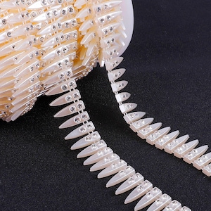 May include: Ivory-colored decorative trim with a repeating pattern of pointed shapes, each adorned with small, sparkling rhinestones. The trim is shown coiled and uncoiled, against a dark background. This trim is suitable for embellishing garments or crafts.