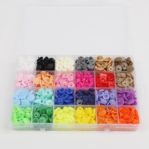 Puede incluir: Una caja de almacenamiento de plástico transparente llena de 18 compartimentos que contienen varios colores de botones a presión de plástico. Los botones están dispuestos en filas y columnas, con cada compartimento que contiene un color diferente.