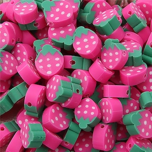 Puede incluir: Un montón de cuentas de plástico en forma de fresa de color rosa y verde. Las cuentas tienen puntos blancos en la parte rosa y una hoja verde en la parte superior.
