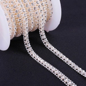 May include: A spool of white rhinestone trim with clear rhinestones. The trim is on a white spool and is unfurled, showing the trim's design.