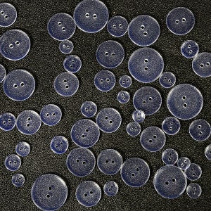 May include: A collection of clear plastic buttons of various sizes, scattered on a black surface. The buttons have two holes each.