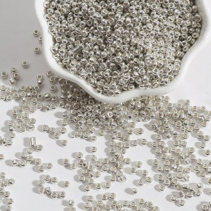 Peut inclure: Un bol blanc rempli de perles de rocaille argentées. Les perles sont éparpillées sur une surface blanche.