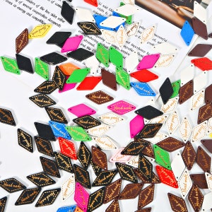 Könnte beinhalten: Eine Sammlung bunter, rautenförmiger Holzetiketten mit dem Text "Hand Made" in goldener Schrift. Die Etiketten sind perfekt, um handgefertigten Produkten, Geschenken und Kleidung eine handgemachte Note zu verleihen.