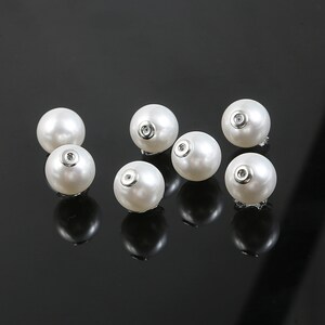 Puede incluir: Ocho botones de nácar blanco con un agujero central de metal plateado. Los botones están dispuestos de forma dispersa sobre una superficie negra.