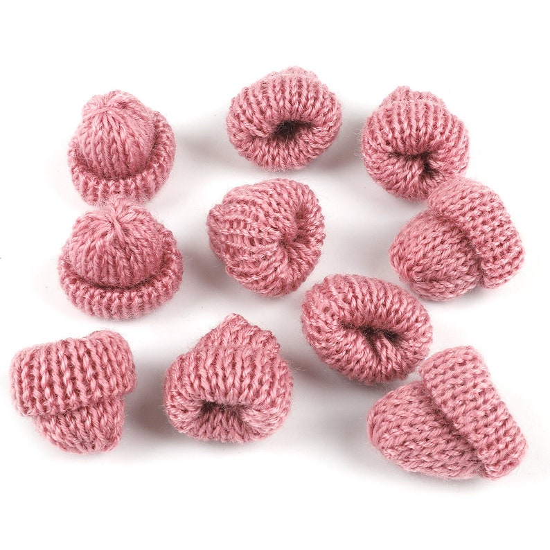 May include: Ten pink knitted mini hats, each with a folded brim, are arranged on a white surface. The hats are approximately 1 inch in diameter.