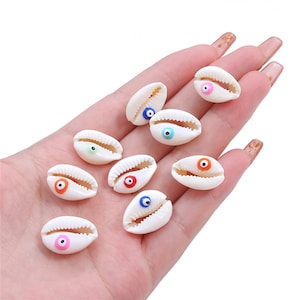 May include: A collection of white seashells with colourful evil eye charms. The shells are oval shaped and have a smooth, polished finish. The evil eyes are made of glass and come in a variety of colours, including blue, green, orange, pink, and red.