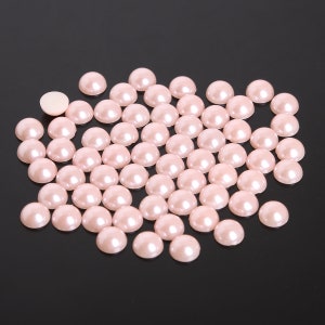 Puede incluir: Un conjunto de perlas de imitación de color rosa claro, de forma semiredonda. Las perlas tienen un acabado brillante y están dispersas sobre una superficie oscura. Las perlas rosas son ideales para manualidades o joyería.