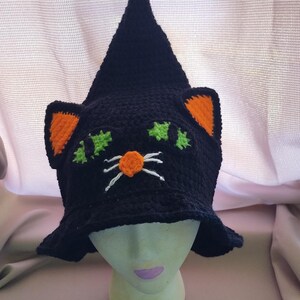 May include: A black crocheted bucket hat shaped like a cat with a pointed top. The hat features two orange inner ears, two bright green eyes with black pupils, and an orange nose with white embroidered whiskers centered on the front panel.