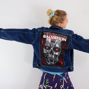 Può includere: Una giacca di jeans blu con una grafica di Terminator Salvation sul retro. La grafica presenta un teschio con occhi rossi e il testo "Terminator Salvation".