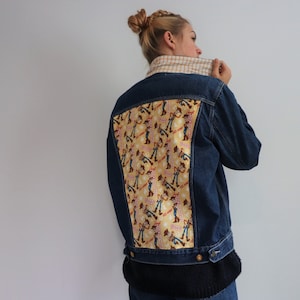 Può includere: Una giacca di jeans blu con una toppa di tessuto giallo e marrone sul retro. La toppa presenta un motivo ripetuto di cowboy dei cartoni animati.