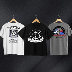 May include: Three t-shirts with different designs. The first t-shirt is white with a penguin illustration and the text "Teamguin of the Month". The second t-shirt is black with a white anchor and the text "Jungle Jim's Adventure Tours". The third t-shirt is gray with a blue truck and the text "The American Towing & Recovery Institute".