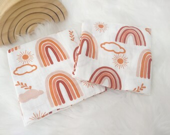 May include: Two white muslin baby burp cloths with a rainbow, sun, and cloud pattern in shades of orange, pink, and brown.