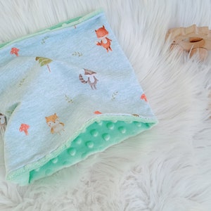 Puede incluir: Una manta para bebé de color azul claro y verde con estampado de animales del bosque. La manta está hecha de un tejido suave y afelpado, ideal para acurrucarse.