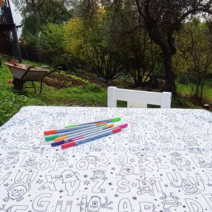 May include: A white tablecloth with a black and white pattern of letters and monsters. There are several colorful markers on the tablecloth.