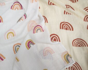 May include: Two white muslin swaddles with a rainbow pattern. One swaddle has a multi-colored rainbow design, while the other has a solid rust-colored rainbow design.