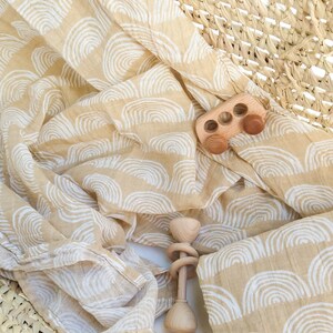 May include: A beige muslin swaddle blanket with a white rainbow pattern. The blanket is folded and draped over a woven basket. Two wooden toys, a car and a rattle, are on top of the blanket.