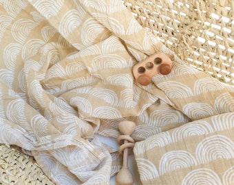 May include: A beige muslin swaddle blanket with a white rainbow pattern. The blanket is folded and draped over a woven basket. Two wooden toys, a car and a rattle, are on top of the blanket.