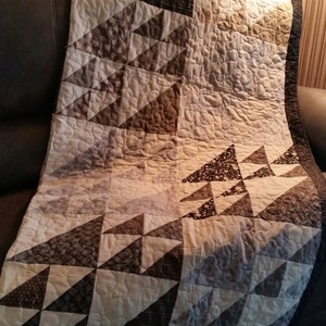 May include: A handmade quilt with a geometric triangle pattern in shades of brown, cream, and black. The quilt has a quilted texture and a dark brown border. The triangles are arranged in rows, creating a modern, abstract design.