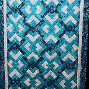 Op de afbeelding: Een handgemaakte quilt met een geometrisch patroon in tinten teal, lichtblauw en wit. Het ontwerp bevat in elkaar grijpende vormen, waardoor een visueel aantrekkelijke en moderne esthetiek ontstaat. De quilt is omrand met een bloemenpatroon.