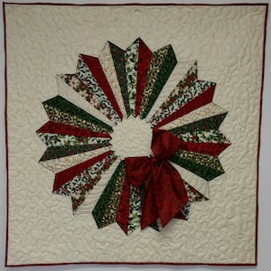 May include: A white quilted fabric square with a red and green Christmas wreath design. The wreath is made of fabric strips with a red bow in the center.