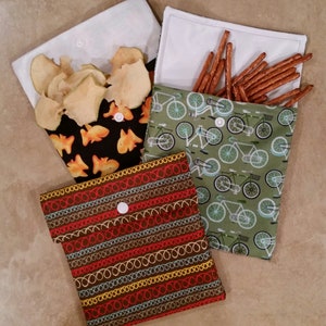 May include: Three reusable snack bags with different patterns. One bag has a black background with orange fish, another has a green background with bicycles, and the last one has a brown background with colorful stripes.