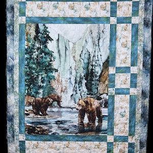 Puede incluir: Un edredón hecho a mano con un paisaje de montaña con osos pardos cerca del agua. El edredón tiene un borde azul y blanco con un diseño central rectangular. Los osos son el punto focal, sobre un fondo de árboles y una cascada.