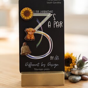 May include: A black display card featuring three pairs of earrings with titanium posts. The card displays the text 3 PAIR COMBINATIONS! 3'S A PAIR BY Different by Design. The earrings feature a sunflower, a highland cow, and a bell design.