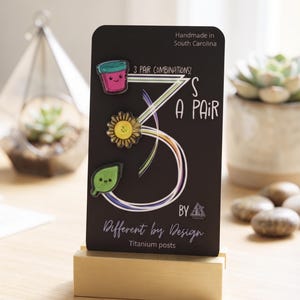May include: A black display card featuring three enamel pins: a pink cup, a yellow sunflower, and a green leaf. The card text reads 3 PAIR COMBINATIONS! 3S A PAIR BY Different by Design, Handmade in South Carolina, and Titanium posts. The card sits in a light wood base.
