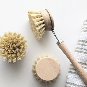 May include: Three natural bristle dish brushes with wooden handles. One brush has a long wooden handle with a metal loop for hanging. The brushes are on a white surface with a striped towel.