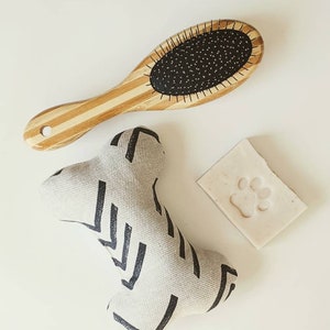 May include: A set of dog grooming supplies including a wooden brush with black bristles, a white and black patterned dog toy shaped like a bone, and a white bar of soap with a paw print.