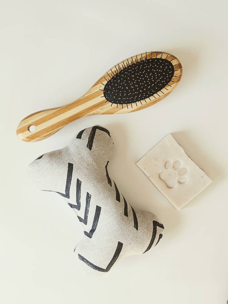 May include: A set of dog grooming supplies including a wooden brush with black bristles, a white and black patterned dog toy shaped like a bone, and a white bar of soap with a paw print.