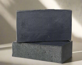 May include: Two bars of charcoal soap stacked on top of each other. The soap is dark grey and has a rough texture.