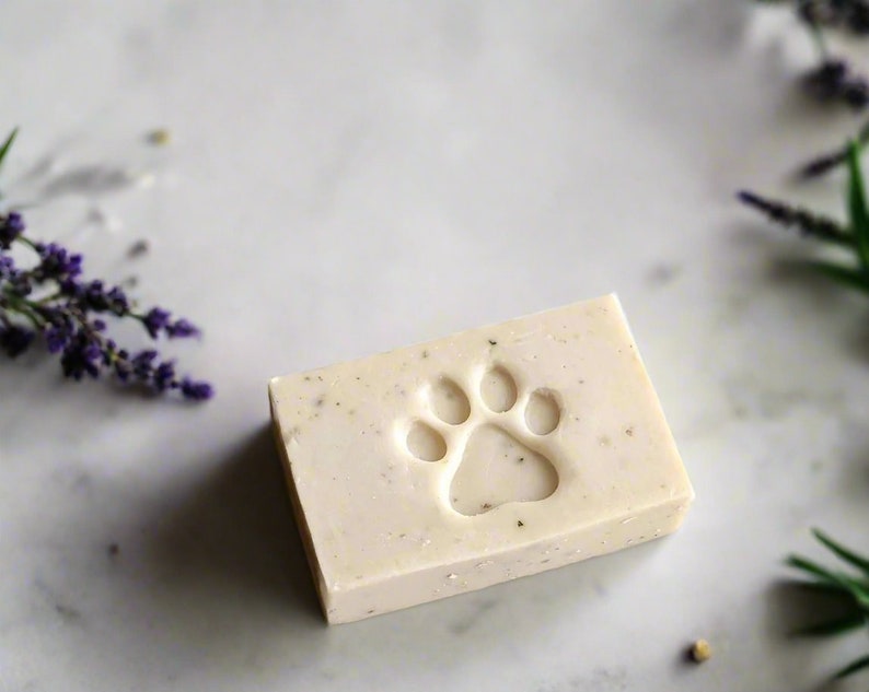May include: A white bar of soap with a paw print design. The soap is on a white marble surface with lavender flowers in the background.