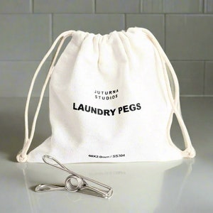 May include: A white drawstring bag with the text "JUTURNA STUDIOS LAUNDRY PEGS 60X2.0mm/SS304" printed on it. A silver metal laundry peg is in front of the bag.