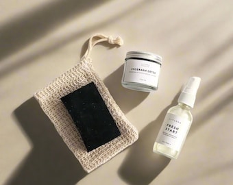 May include: A set of natural deodorant products in a beige and white color scheme. The set includes a small glass jar labeled "Underarm Detox", a clear spray bottle labeled "Fresh Start", and a beige mesh bag containing a black bar of soap.