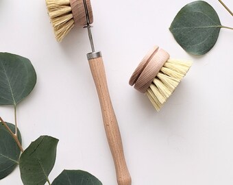 May include: Two wooden dish brushes with natural bristles. The brush on the left has a long wooden handle with a metal loop at the end. The brush on the right is smaller and has a shorter handle. Both brushes are on a white background with green eucalyptus leaves.