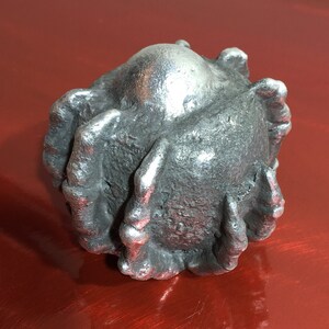 May include: A silver-coloured, round, decorative object with a textured surface and a smooth, round top. The object has a series of raised, claw-like protrusions around the perimeter.