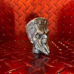 Peut inclure: Une tête sculptée argentée avec une surface texturée, possiblement un bouchon de bouteille ou un objet décoratif. La tête a une apparence vieillie et détaillée, avec un nez et une barbe proéminents. Le fond est une surface rouge en losange.