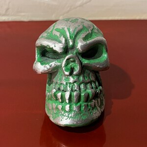 May include: A green and silver skull figurine with a menacing expression. The skull has a detailed face with sharp teeth and a textured surface.