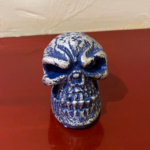 Peut inclure: Une figurine de crâne bleu et argenté. Le crâne présente un design détaillé avec des accents argentés sur le front, les joues et la mâchoire. La figurine est faite d'un matériau solide et repose sur une surface rouge.
