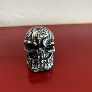 May include: A small, silver skull figurine with a detailed, textured surface. The skull has a menacing expression with sharp teeth and deep eye sockets.