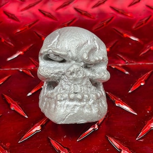 May include: A silver skull sculpture with a detailed, textured surface. The skull has a menacing expression with a furrowed brow and a wide, toothy grin.