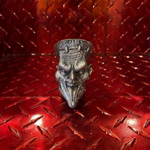 May include: A silver-coloured, detailed sculpture of a monster's head, possibly Frankenstein's monster, with a textured surface. The sculpture has a stern expression and is set against a red diamond plate background.