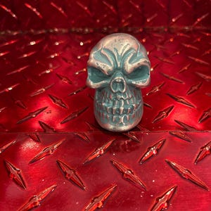 May include: A teal-colored skull figurine with silver accents. The skull has detailed facial features and is set against a red, diamond-plate background. The figurine is a decorative item.