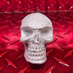 May include: A silver skull sculpture with a detailed face and textured surface. The skull is sitting on a red diamond plate background.