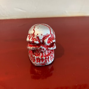 May include: A small, silver skull figurine with red paint splatters. The skull has a detailed face with a menacing expression.