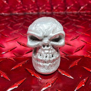 May include: A silver skull-shaped gear shift knob with a detailed design. The knob is sitting on a red diamond plate surface.