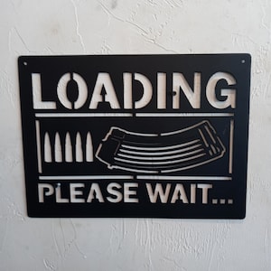 May include: Black metal sign with the words "LOADING" and "PLEASE WAIT..." in white. The sign features a stylized image of a magazine and bullets.