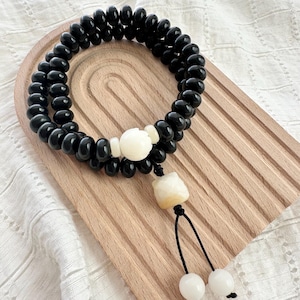 May include: Black beaded bracelet with a white floral accent and a decorative tassel. The bracelet is displayed on a wooden tray with a white fabric background. The bracelet is made of small, round beads.
